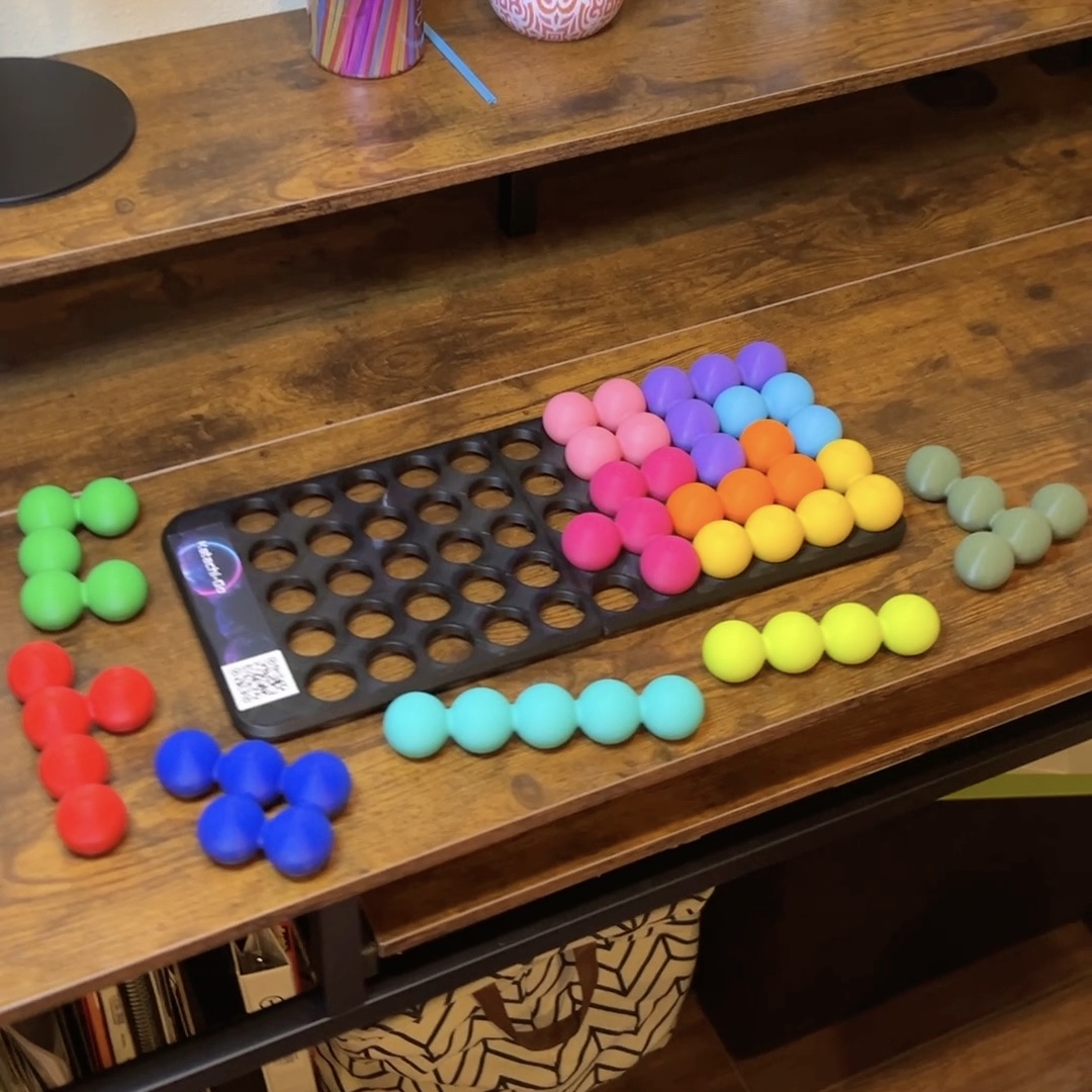 A spatial logic puzzle tray half filled with colorful fused-ball pieces, with the remaining pieces laid out beside it on a wooden desk