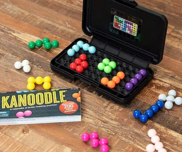An open Kanoodle case with pieces in its tray, and more pieces and the challenge booklet on a wooden table